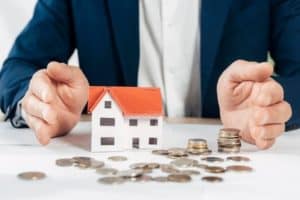 A person in a blue suit sits at a desk, with their hands held up on either side of a small model house with a red roof. Scattered around the house are various stacks and piles of coins, suggesting themes of property, assets, and financial protection or risk.