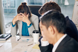 Three people are seated around a conference table, appearing stressed and overwhelmed. One woman has her face in her hands, while the two men next to her also look troubled. Papers with charts and a coffee cup are on the table, suggesting a difficult business meeting or financial discussion.