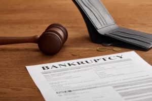A bankruptcy form is prominently displayed on a wooden desk, with a wooden gavel and an open, empty wallet beside it. A few coins are scattered near the wallet, emphasizing financial hardship and the legal process of bankruptcy.