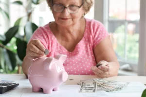 An older woman wearing glasses and a pink shirt is seen actively putting coins into a pink piggy bank. On the table in front of her are scattered banknotes and coins, along with a calculator and papers, symbolizing retirement savings, financial planning, or managing money during difficult times.