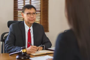 A male attorney wearing glasses, a suit, and a red tie is seated at a desk, looking towards a client (whose back is to the camera). He is holding a pen and taking notes in a notebook, with a gavel visible on the desk, indicating a legal consultation or discussion about significant financial decisions like filing for bankruptcy.
