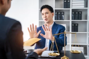A man in a blue suit sits at a desk, holding up both hands in a "stop" gesture towards another person who is holding out a yellow envelope. A set of golden scales of justice is visible on the desk, symbolizing legal boundaries and the protection a bankruptcy lawyer offers against creditor harassment.