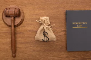 A top-down view of a wooden desk featuring a wooden gavel on the left, a small burlap money bag with a dollar sign in the center, and a dark legal book titled "BANKRUPTCY LAW" on the right. This arrangement symbolizes the legal aspects of bankruptcy and debt relief.