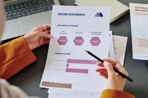 A person's hands are holding an "INCOME STATEMENTS" document, pointing with a pen at "COMPANY EXPENSES" on a dark desk. The document details revenue sources and expenses with bar graphs and figures. A laptop and other financial papers are visible, suggesting financial analysis, budgeting, or reviewing income for bankruptcy eligibility.